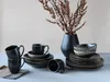 A collection of dark-colored ceramic dishes and cups are arranged on a light-colored surface, with a vase of dried flowers in the background. Produktbild-Vorschau