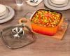 A square orange pot filled with food sits on a wooden cutting board, next to its glass lid. The scene is set on a wooden table with plates and cutlery in the background. Produktbild-Vorschau