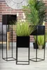 Three black metal plant stands with artificial plants are arranged in a room with a brick wall and a large clock in the background. Produktbild-Vorschau
