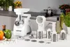 A white Zelmer meat grinder and its accessories are displayed on a white surface with a kitchen counter in the background. Produktbild-Vorschau