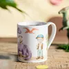 A mug with dinosaur illustrations is placed on a wooden surface, with a green plant and a toy dinosaur in the background. Produktbild-Vorschau