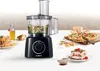 A Bosch food processor is shown on a white surface with various food items, including a salad, bread, a quiche, and a dessert, suggesting its versatility in food preparation. Produktbild-Vorschau