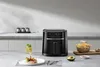 An air fryer is displayed on a kitchen counter with food and a drink. Produktbild-Vorschau