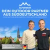 Two men are standing in front of a cityscape with a prominent church tower. The background is a bright blue sky. Produktbild-Vorschau