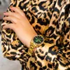 A close-up shot of a woman's arm wearing a gold watch with a green face, against a leopard print fabric. Voir les images du produit