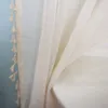 A close-up of a cream-colored curtain with tassels, draped over a white surface, with a light blue wall in the background. Produktbild-Vorschau