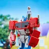 A toy Optimus Prime robot with a small human figure standing on its head, set against a blurred outdoor background of trees and a house. Podgląd zdjęcia produktu
