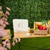 A white money box and crates of fresh vegetables are displayed on a wooden table in front of a green ivy wall. Náhled obrázku produktu