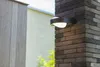 An outdoor wall light mounted on a brick pillar next to a wooden fence. Produktbild-Vorschau