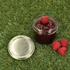 A jar of raspberry jam with raspberries and its lid on a grassy surface. Podgląd zdjęcia produktu