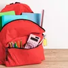 A red backpack filled with school supplies, including notebooks, pencils, a calculator, and a ruler, is shown on a wooden surface. Produktbild-Vorschau