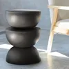 A dark gray, three-tiered stool or side table is placed on a concrete floor next to a light-colored armchair. Sunlight streams in from the right, casting shadows. Produktbild-Vorschau