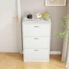 A white shoe cabinet with three drawers is shown in a room next to a white door and a window with curtains. Náhľad obrázku produktu