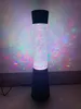 A tall, cylindrical lamp with a black base and top, filled with colorful glitter and illuminated from within. Anteprima dell’immagine prodotto