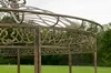 A close-up view of a decorative metal gazebo with intricate floral designs, set against a backdrop of green grass and trees. Anteprima dell’immagine prodotto