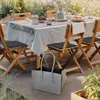 An outdoor dining table set up for a meal, with chairs, a tablecloth, and food items on the table. A grey felt shopping bag is placed on the wooden deck in the foreground. Produktbild-Vorschau