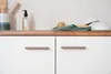 A close-up view of a white kitchen cabinet with two drawers, each featuring a silver handle. On the wooden countertop above the cabinets, there are kitchen items including a bottle of soap, a green cloth, a wooden brush, and stacked white bowls. Produktbild-Vorschau