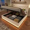 A white upholstered bed with its storage compartment open, revealing neatly folded linens and towels. The bed has LED lighting around its base and a tufted headboard. A window with a city view at night is visible in the background. Náhled obrázku produktu