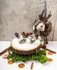 A Christmas decoration display featuring a reindeer figure, pine cones, and glass ornaments on a wooden slice, surrounded by pine branches, cinnamon sticks, and dried orange slices. Podgląd zdjęcia produktu