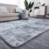 A fluffy gray rug is placed in a living room setting in front of a sofa. Decorative pillows and a throw blanket are on the sofa, and a plant and a cabinet are in the background. Produktbild-Vorschau
