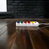 A white power strip with colored outlets sits on a dark wooden floor in front of a dark brick wall. Produktbild-Vorschau