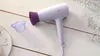 A white hairdryer with a purple nozzle and a detached purple diffuser attachment is placed on a white wooden surface, with delicate white flowers in the background. Podgląd zdjęcia produktu