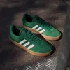 A pair of green Adidas VL Court 3.0 sneakers with white stripes and tan soles are displayed on a textured gray surface. Produktbild-Vorschau