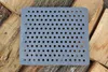 A gray metal grate with many holes is placed on top of stacked pieces of wood. Voir les images du produit