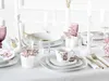 A beautifully set dining table with white porcelain plates, cups, and glassware, adorned with delicate pink flowers and gold cutlery. Produktbild-Vorschau