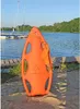 A bright orange rescue buoy is placed on a sandy beach next to a wooden dock, with a calm lake and sailboats in the background. Produktbild-Vorschau