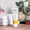 A tube of Medela PureLan Lanolin cream is displayed on a textured surface, with a stack of white towels and a blurred background of bathroom items. Náhled obrázku produktu