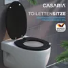A black toilet seat is shown on a white toilet against a tiled wall background, with some greenery in the foreground and background. Anteprima dell’immagine prodotto