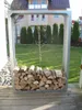 A metal firewood rack filled with logs is placed on a wooden deck in front of a grassy yard and a house. Produktbild-Vorschau