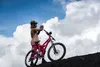 A person with a mountain bike on a dark, rocky surface with a cloudy blue sky in the background. Anteprima dell’immagine prodotto