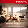 A modern living room with a large window overlooking a lake, featuring a sofa, armchair, and a coffee table. The floor has visible heating elements. Produktbild-Vorschau