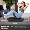 A woman is performing a sit-up exercise on a black vibration plate with purple accents, in a living room setting. Produktbild-Vorschau