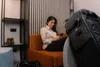 A woman is sitting on an orange couch in a room, looking at her phone. A black bag with a tracking device is in the foreground. Podgląd zdjęcia produktu