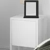 A white nightstand with a black e-reader on top, placed next to a bed. Produktbild-Vorschau