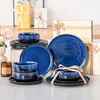 A collection of blue and black ceramic dinnerware, including plates and bowls, arranged on a table with gift boxes in the background. Produktbild-Vorschau