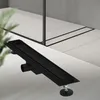 A black linear shower drain is shown on a tiled floor, with a glass shower enclosure in the background. Náhled obrázku produktu