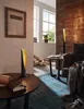 A cozy living room scene with two modern table lamps, a book, and a rolled-up paper on a wooden table, with a fireplace and windows in the background. Anteprima dell’immagine prodotto