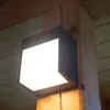 A modern black LED outdoor wall light with a white illuminated panel is mounted on a wooden beam, with wooden planks visible in the background. Podgląd zdjęcia produktu
