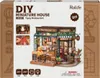 The image shows the packaging of a DIY miniature house kit, specifically a tipsy restaurant model. The packaging displays the product name, brand, and a detailed image of the assembled miniature. Voir les images du produit
