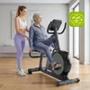 An older woman is exercising on a recumbent exercise bike, with a younger woman assisting her. The scene is indoors, likely a home gym or fitness studio. Produktbild-Vorschau