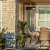 A white outdoor sensor is mounted on a stone wall next to a door and windows, with patio furniture and plants visible. Podgląd zdjęcia produktu
