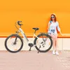 A woman is sitting on a bench next to an electric bicycle, looking at her phone. Produktbild-Vorschau