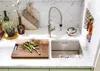 A kitchen sink area with a faucet, cutting board with green beans, and various kitchen accessories. Podgląd zdjęcia produktu