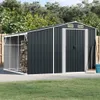 A dark gray metal shed with a lean-to attached, storing firewood. The shed is situated on a concrete pad in a grassy yard with trees in the background. Produktbild-Vorschau