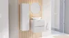 A modern white bathroom vanity with ribbed fronts and a matching tall cabinet, set against a slatted wood panel wall. A round mirror and a white towel complete the scene. Podgląd zdjęcia produktu