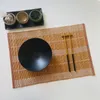 A table setting with a bamboo placemat, a black bowl, and chopsticks. Produktbild-Vorschau
